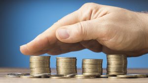 Eine menschliche Hand schützt vier geordnete Stapel mit Goldmünzen auf einem Holztisch vor einem blauen Hintergrund, symbolisch für finanziellen Schutz oder Sparmaßnahmen.