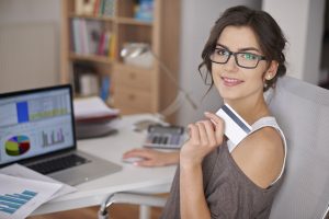 Eine junge Frau mit Brille sitzt an einem Schreibtisch, hält eine Kreditkarte in der Hand und lächelt in die Kamera. Auf dem Tisch befinden sich ein Laptop mit Finanzdiagrammen auf dem Bildschirm sowie weitere Arbeitsmaterialien. Die Szene symbolisiert moderne Kreditmodelle und deren Nutzung durch digitale Finanzanwendungen im privaten und beruflichen Alltag.