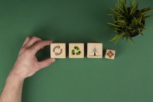 Eine Hand arrangiert vier Holzklötze mit Symbolen, die Konzepte der Kreislaufwirtschaft darstellen: ein Recycling-Symbol, ein Windrad für erneuerbare Energien und weitere Nachhaltigkeitszeichen. Daneben steht ein kleiner Topf mit einer Pflanze auf einem grünen Hintergrund, der die Verbindung zur Umwelt und ökologischen Verantwortung unterstreicht.