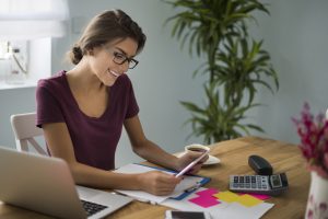 Eine junge Frau arbeitet lächelnd an einem Schreibtisch mit Laptop, Taschenrechner und Unterlagen – sie informiert sich über Steuervorteile und organisiert ihre Finanzen effizient im Homeoffice.
