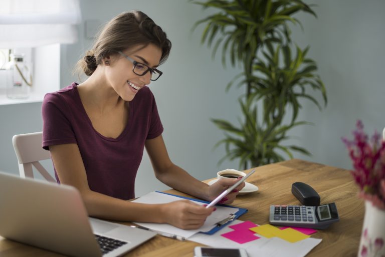 Eine junge Frau arbeitet lächelnd an einem Schreibtisch mit Laptop, Taschenrechner und Unterlagen – sie informiert sich über Steuervorteile und organisiert ihre Finanzen effizient im Homeoffice.