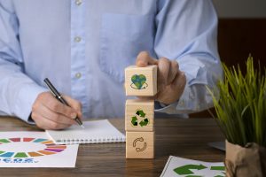 Ein Mann stapelt Holzwürfel mit Recycling- und Umweltsymbolen neben einem Notizblock mit der Agenda 2030 – ein Symbol für nachhaltige Investitionen und strategische Planung im grünen Markt.