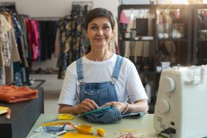 Eine lächelnde Frau mittleren Alters sitzt an einem Tisch in einem Atelier oder Geschäft, näht an einem Kleidungsstück und ist von Nähutensilien umgeben, darunter Schere, Maßband und Nähmaschine. Im Hintergrund hängen verschiedene Kleidungsstücke auf Kleiderständern. Die Szene vermittelt Kreativität und Unternehmergeist, ein Symbol für kleine Investoren, die in handwerkliche oder lokale Geschäftsideen investieren.