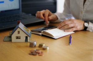 Eine Person berechnet mit einem Taschenrechner und einem Notizbuch ihre Finanzen, während ein Miniaturhaus und Münzen auf dem Tisch liegen – Symbolbild für Immobilieninvestitionen in Deutschland und finanzielle Planung.