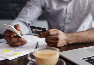 Ein Mann überprüft mit Smartphone und Kreditkarte seine Rechnungen an einem Schreibtisch mit Unterlagen, Laptop und Kaffee – Symbolbild für Online-Zahlungen und Ratenzahlung im Alltag.