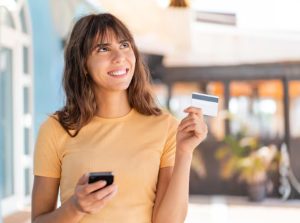 Junge Frau im Freien, die lächelnd ein Smartphone und eine Karte in der Hand hält; die Szene symbolisiert die flexible Nutzung moderner Prepaid-Kreditkarten für sicheres mobiles Bezahlen.